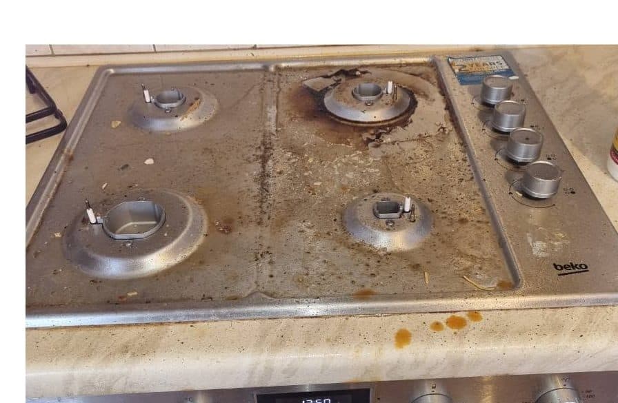 A four-burner stainless steel gas hob with burnt-on brown spill around the burners and along the front trim